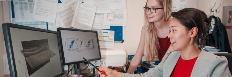 Two PhD students are looking at two computer screens with graphs on one, brain slice scans on the other