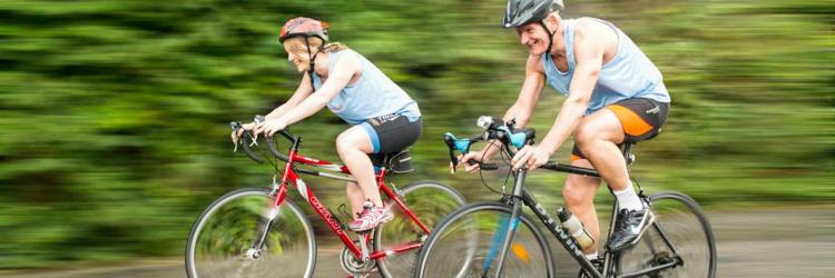 Two people happily cycling fast past camera wearing shorts, shirt and cycling helmets