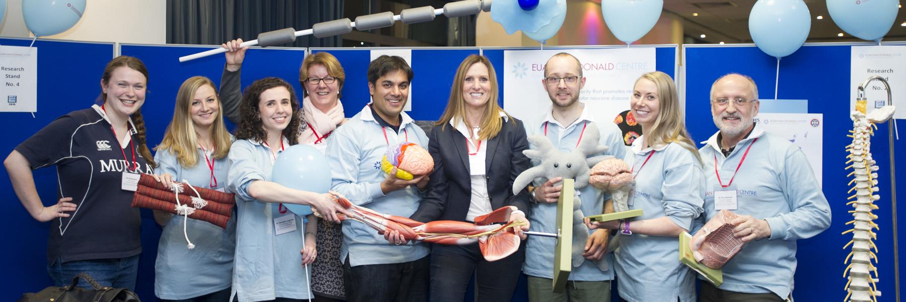 a group of nine Euan MacDonald Centre members at an event, holding props they have used to demonstrate their research