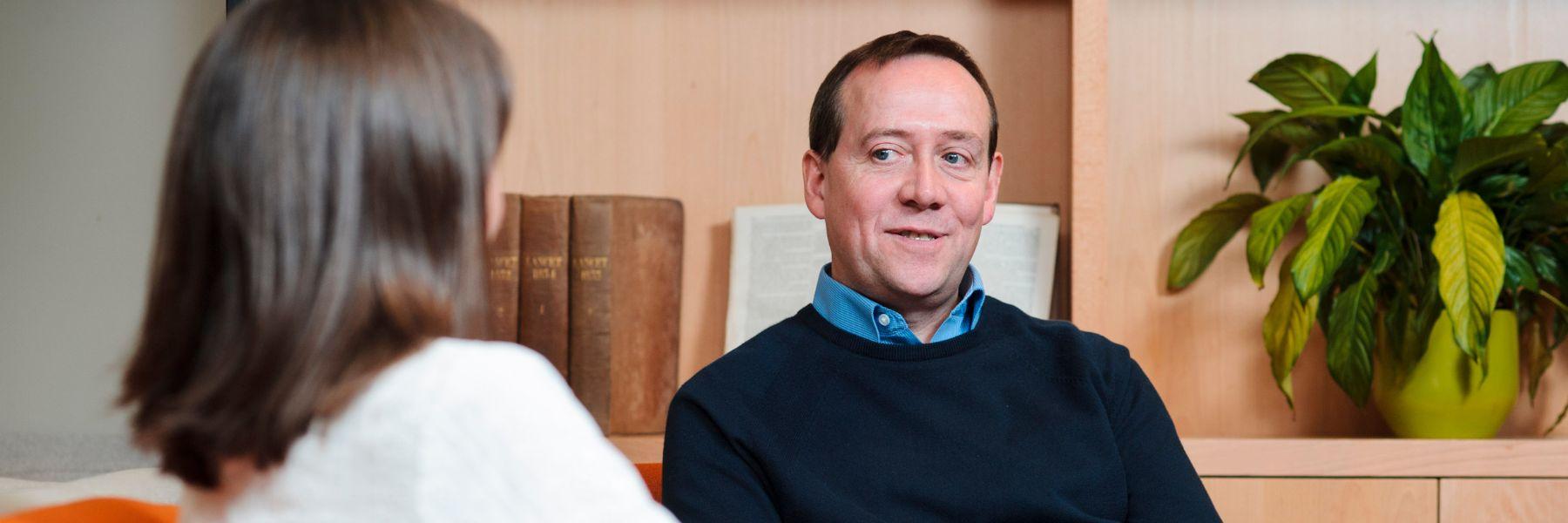 Two people in relaxed discussion in a brightly lit room with a bookcase in the background.
