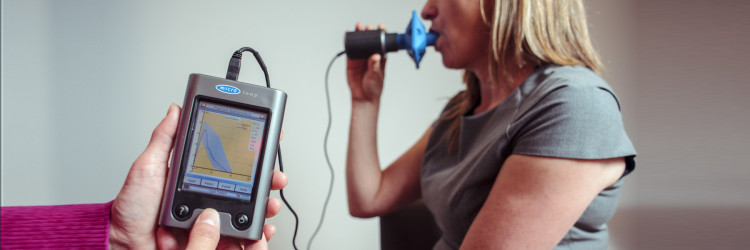 A close up of the screen of a spirometer (an instrument that measures breathing), with an out-of-focus patient in the background doing the test, by blowing into the mouthpiece