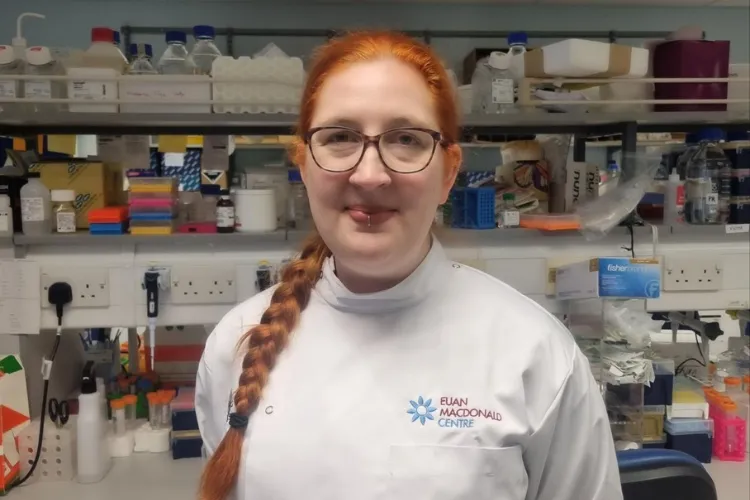 A picture of Denise O'Keefe wearing a Euan MacDonald Centre lab coat the picture is taken with the laboratory as the background.