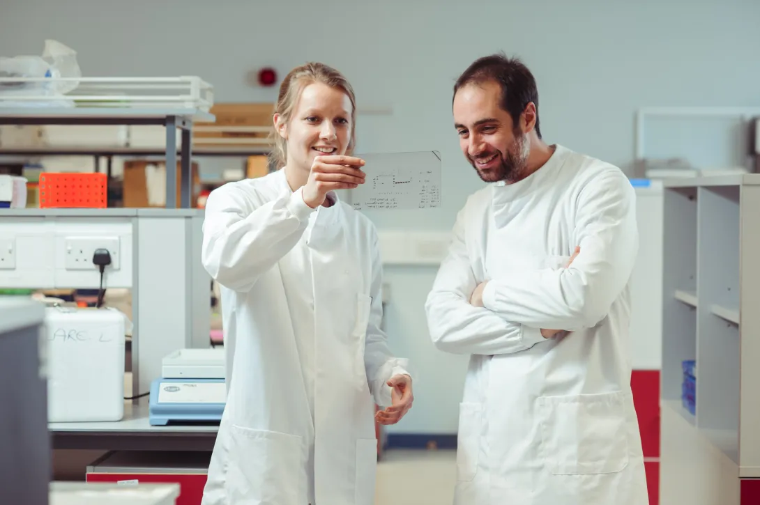 Two PhD students wearing lab coats are smiling as they look at some research data