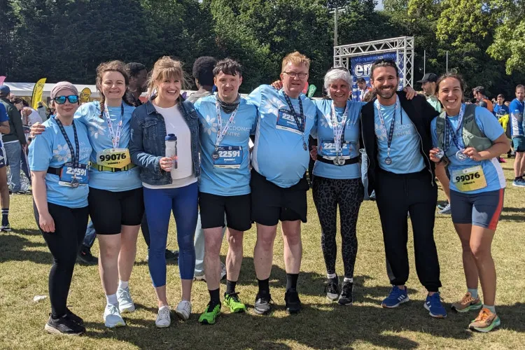 Eight people standing with linked arms outside near the finish line of the Edinburgh marathon relay, wearing light blue Euan MacDonald Centre T-shirts and with medals around their necks